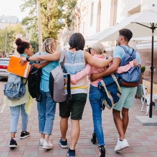 Portrait from back of students with stylish backpacks walking down the street after lectures in university. Tall brunette young man embracing girls while spending time with them outdoot.